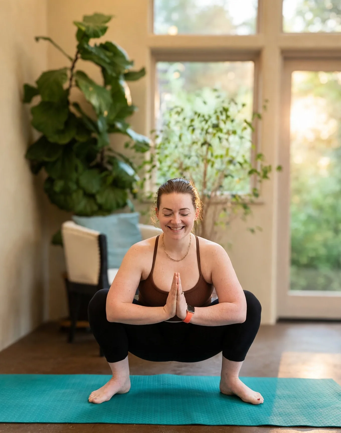 Sara Ahmadi smiling in malasana with prayer hands in a bright room filled with plants